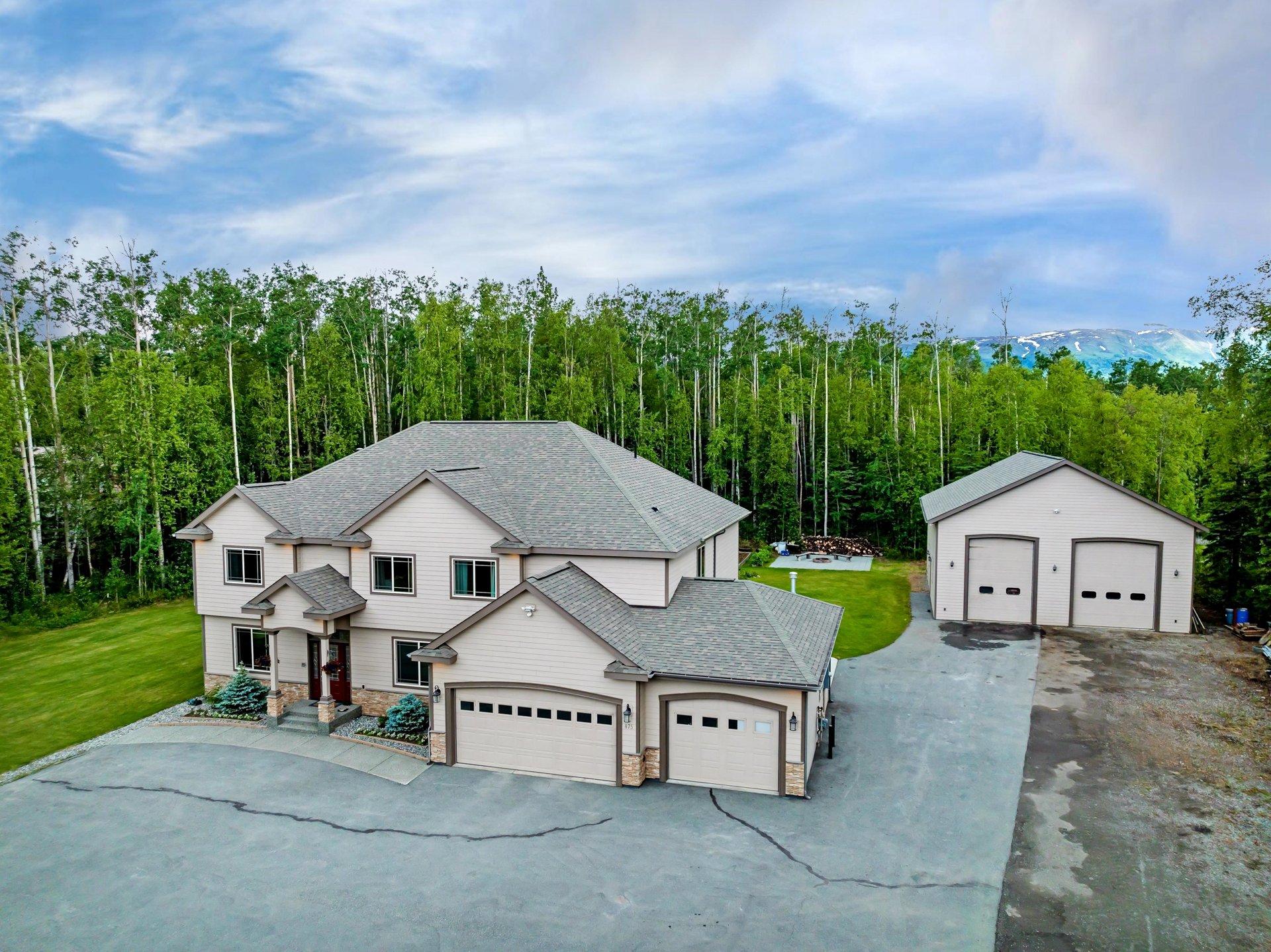 Aerial view of a modern two-story home with a large driveway, two garage doors, and a detached garage, surrounded by green trees with snow-capped mountains in t