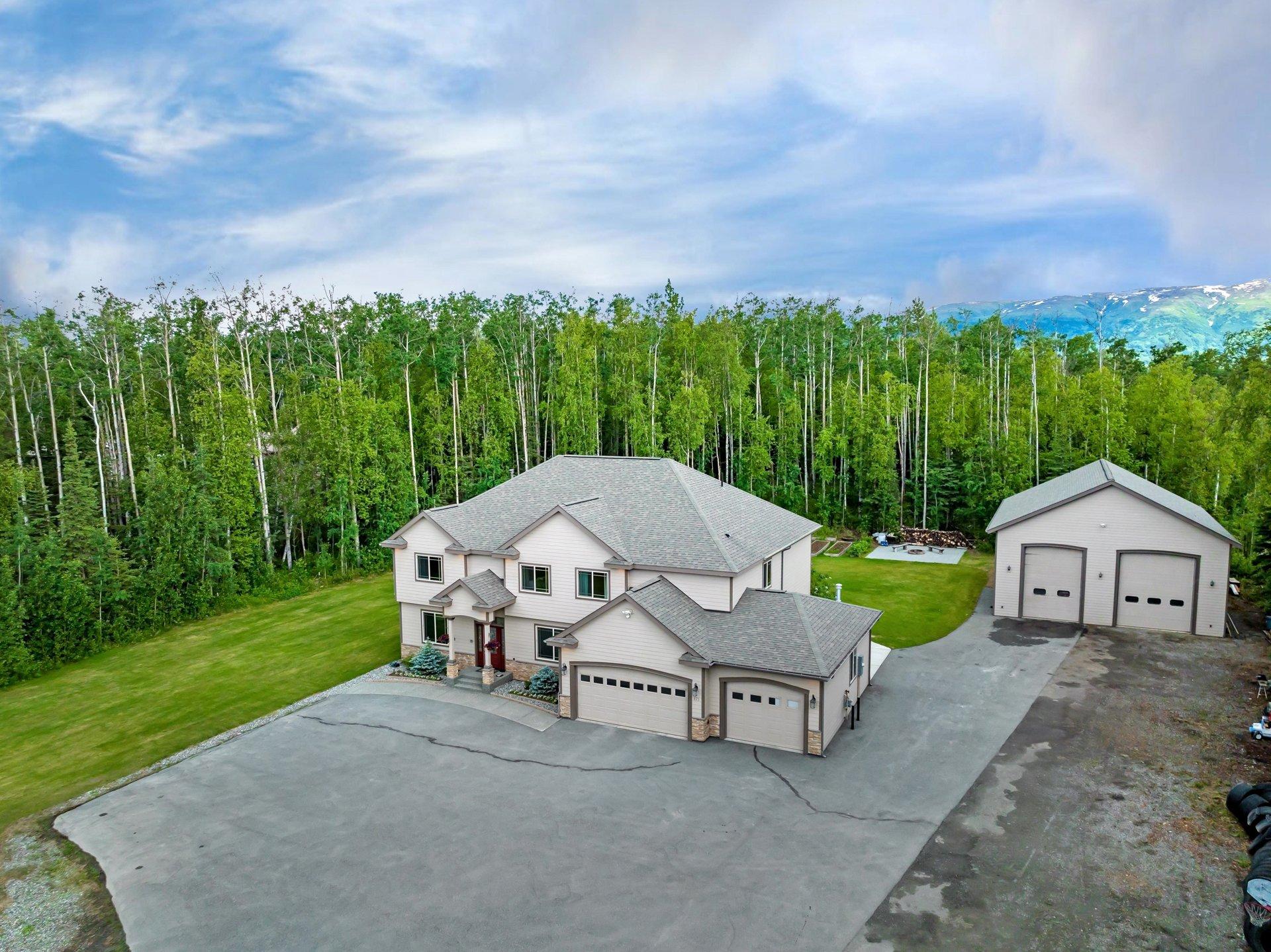 Aerial view of a modern two-story home with a large driveway and attached garage, surrounded by lush green trees and a distant mountain range with snow-capped p