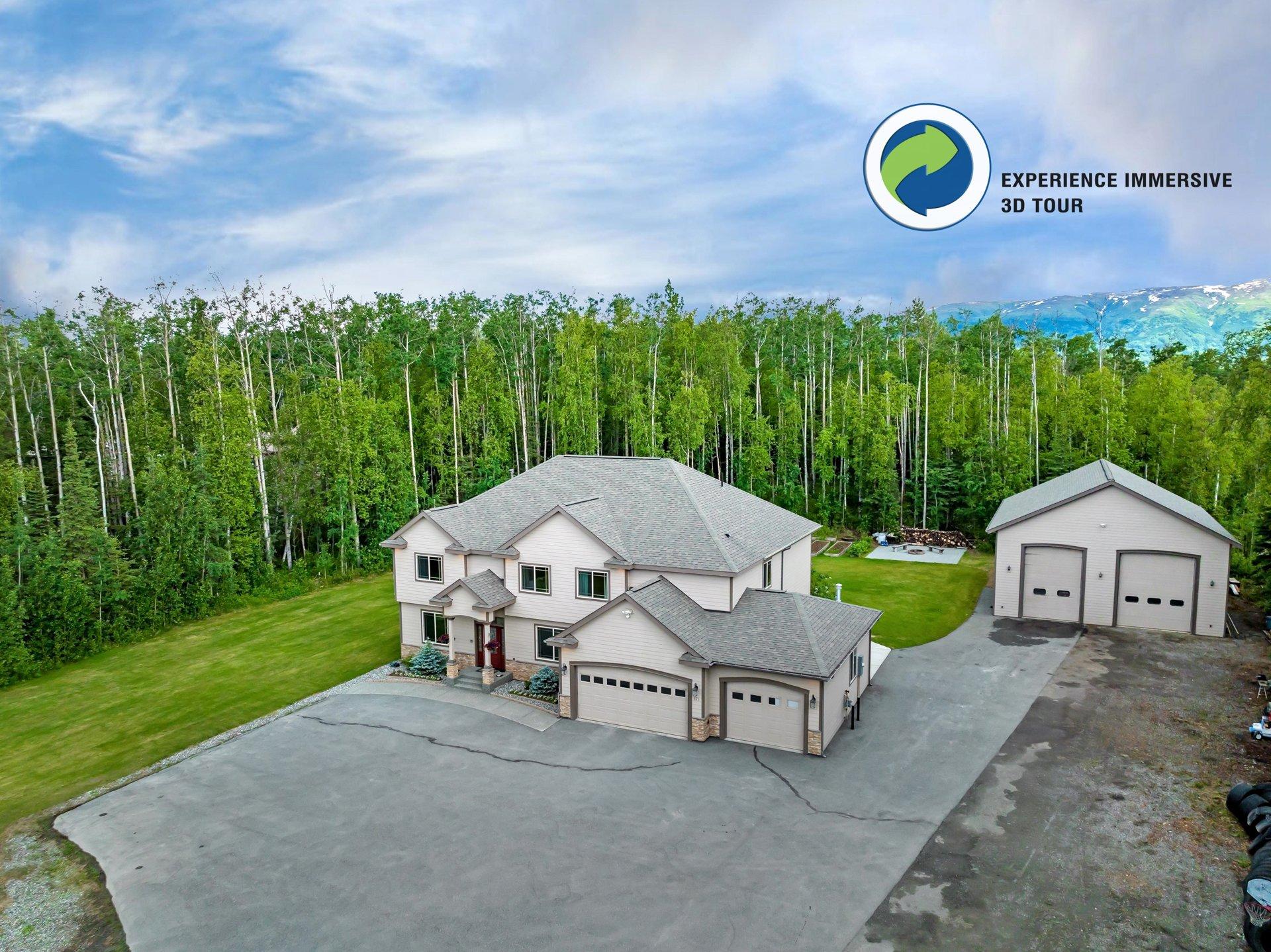 Aerial view of a modern two-story home with a large driveway and attached garage, surrounded by lush green trees and a mountain range in the background with sno
