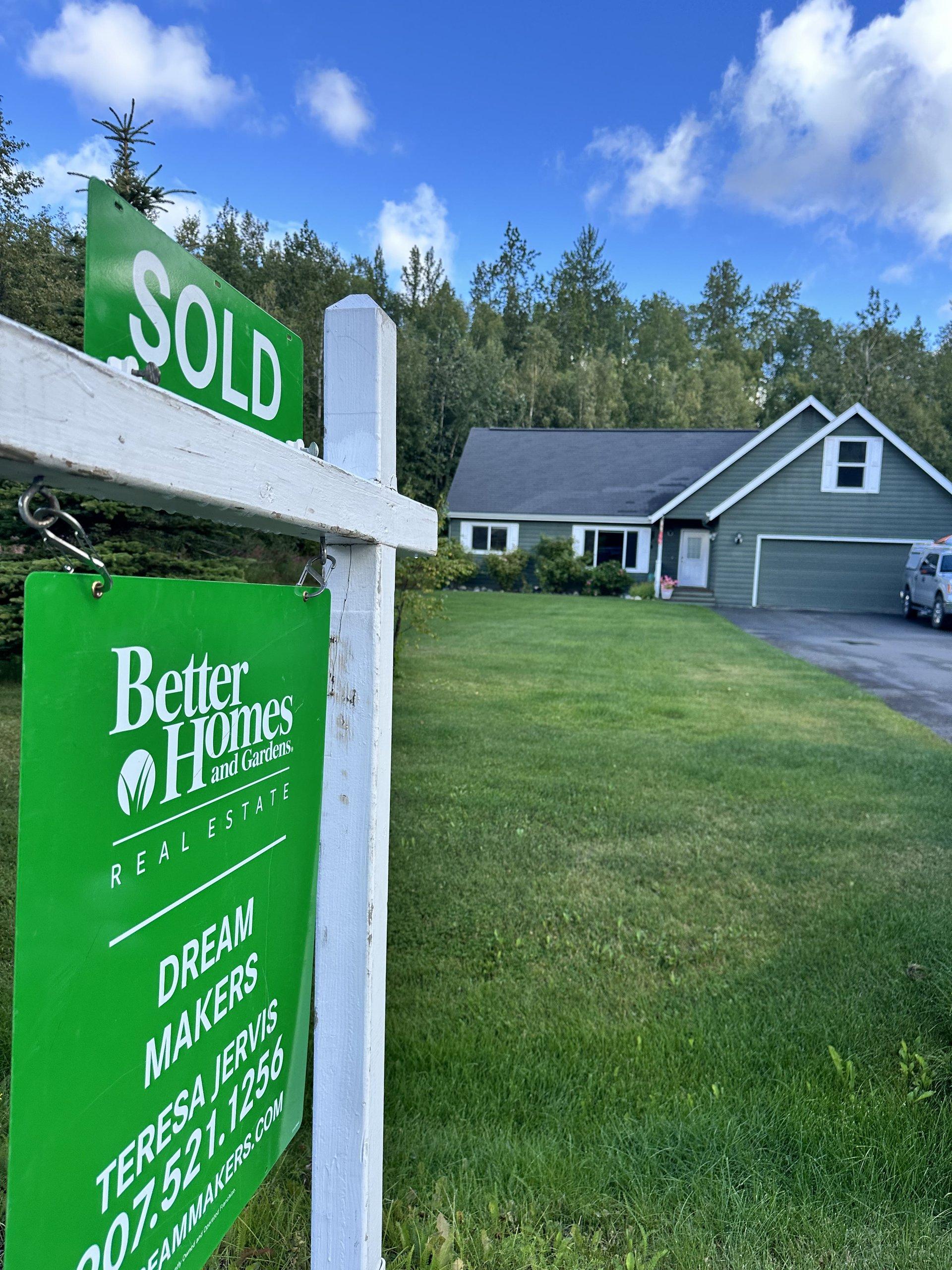 A green 'SOLD' sign for Better Homes and Gardens Real Estate Dream Makers stands in front of a single-family home with a well-maintained lawn, surrounded by tre