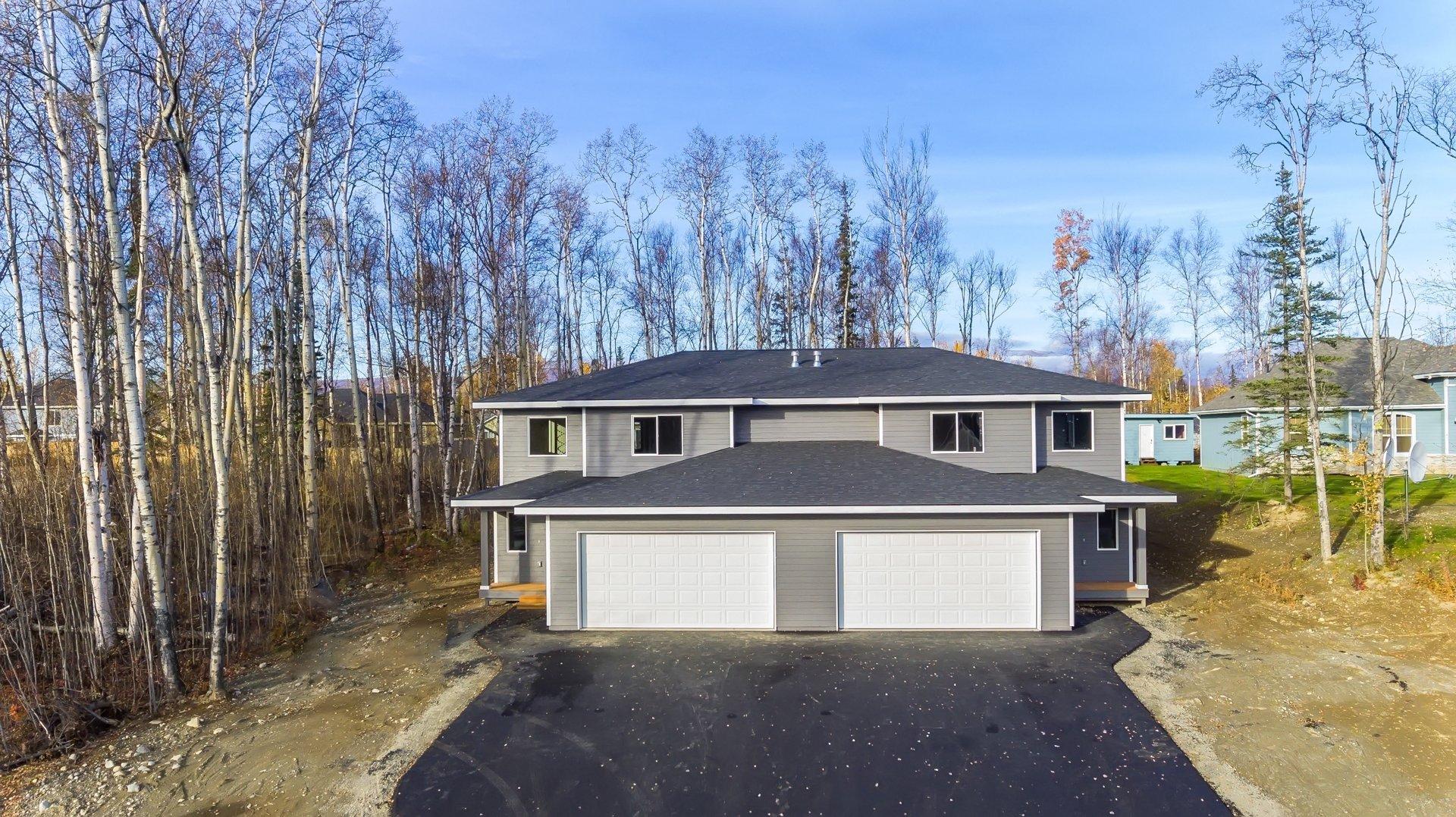 A modern gray two-story house with a double garage, surrounded by bare birch trees and a gravel driveway under a clear blue sky.