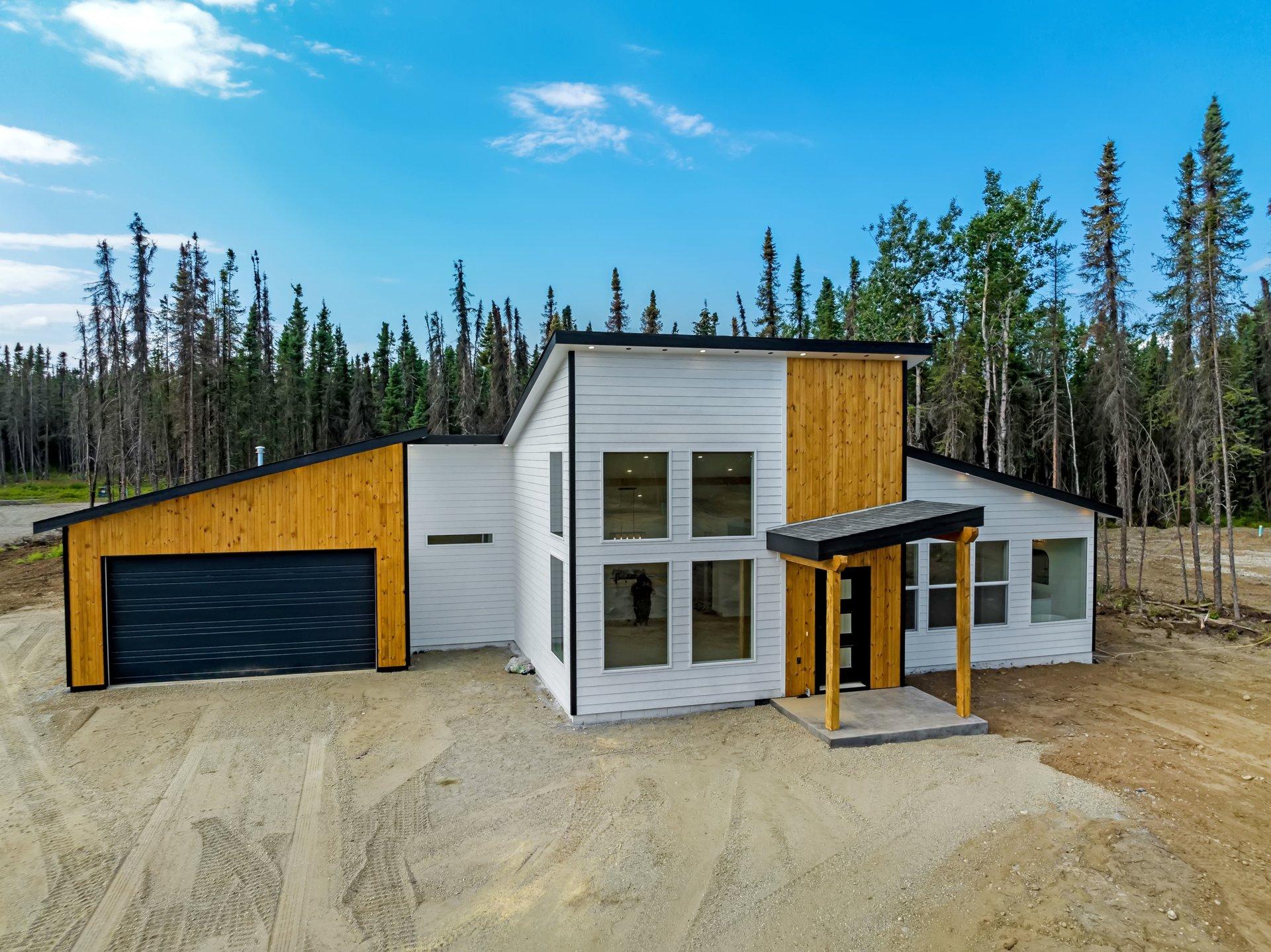 A modern two-story house with white siding and wood accents, featuring large windows and a black garage door, set against a backdrop of tall evergreen trees und