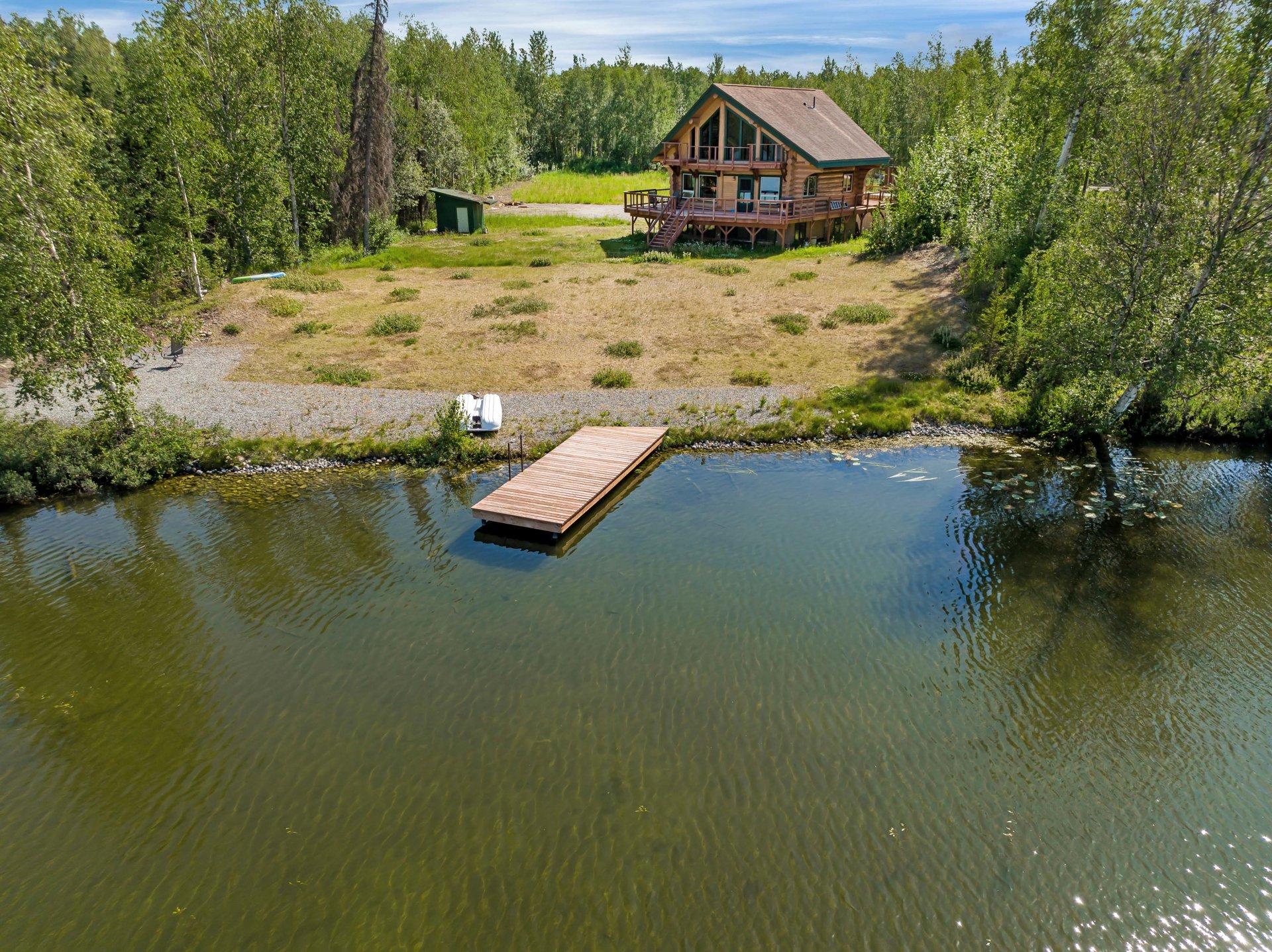 An aerial view of a log cabin on a grassy lot beside a calm body of water with a wooden dock extending into it, surrounded by dense green trees.