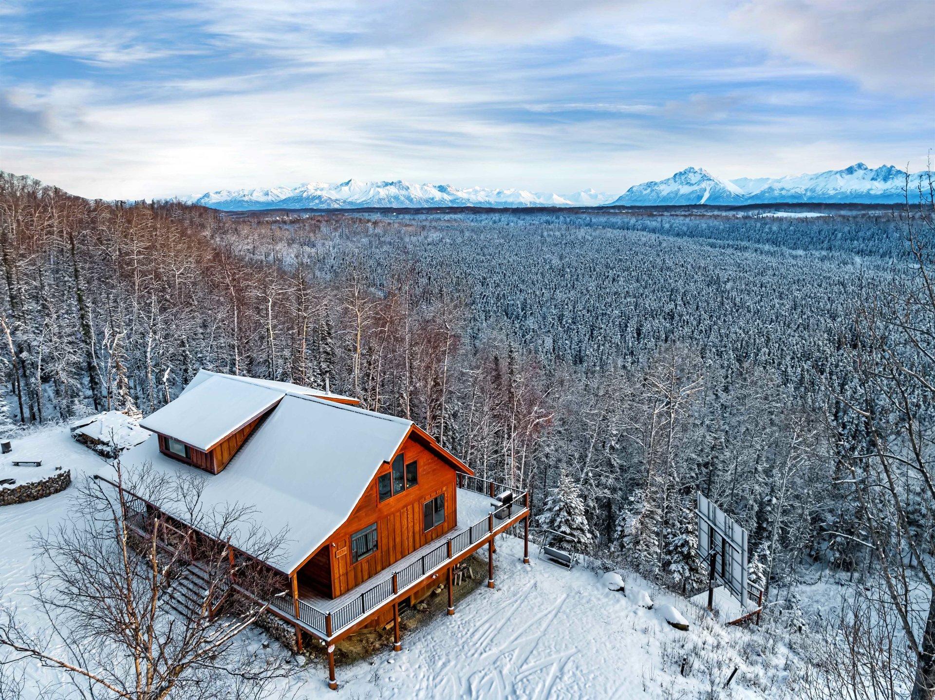 Aerial view of a wooden cabin with a snow-covered roof and deck, nestled on a snowy hillside overlooking a vast, snow-laden forest and distant snow-capped mount
