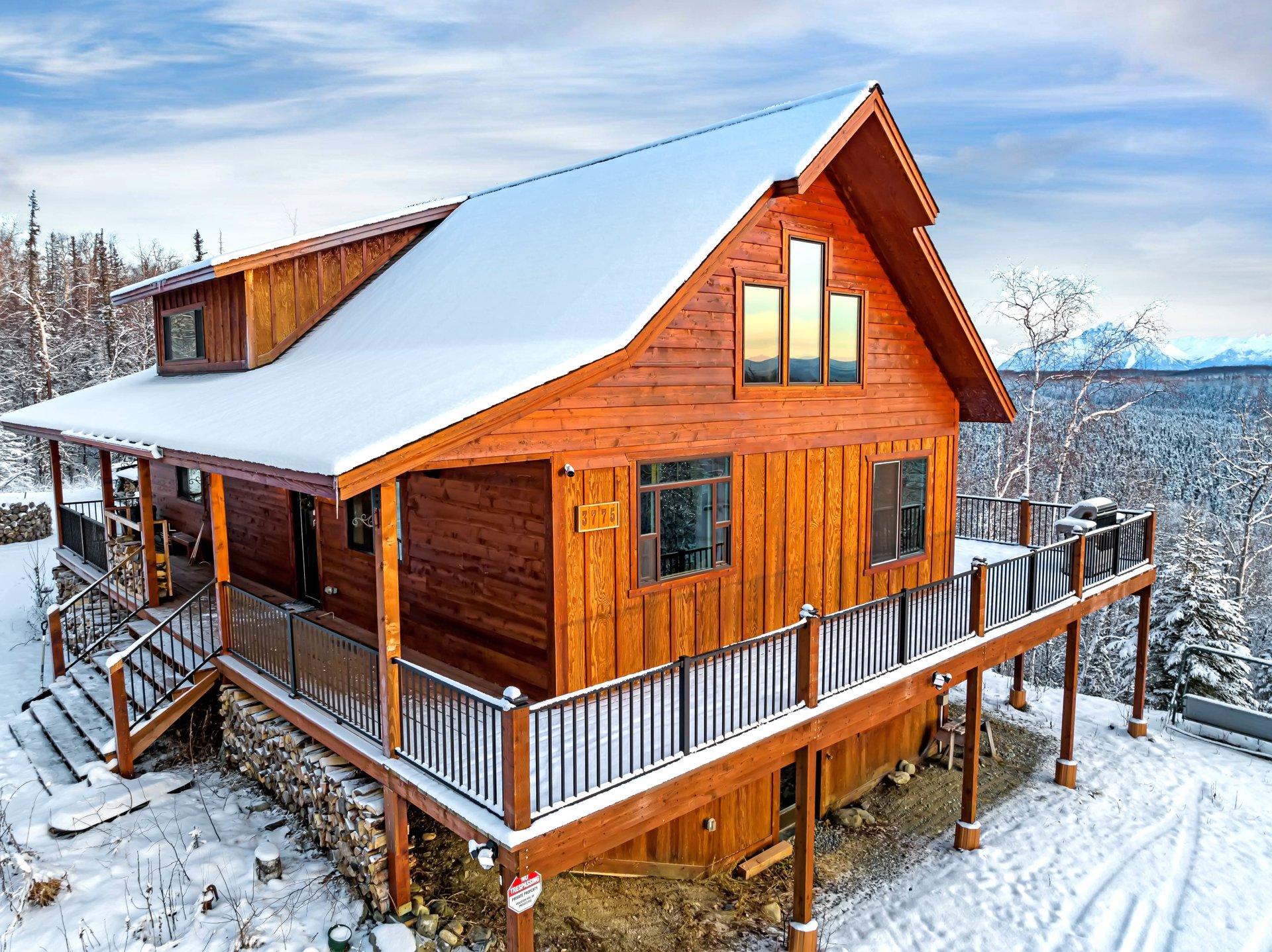 A snow-covered wooden cabin with a large deck and mountain view, set in a winter landscape with trees and distant snow-capped peaks.