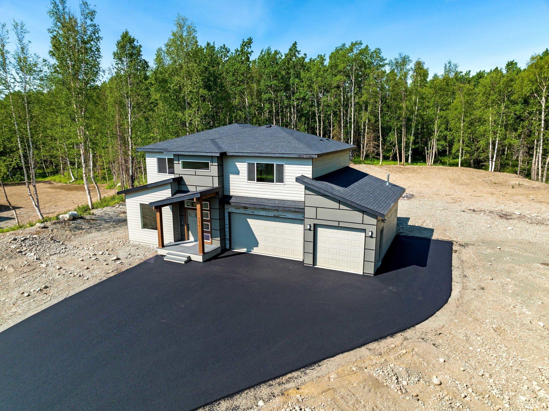 A modern two-story house with a dark roof and light siding sits on a gravel lot with a newly paved asphalt driveway, surrounded by a dense forest of tall birch