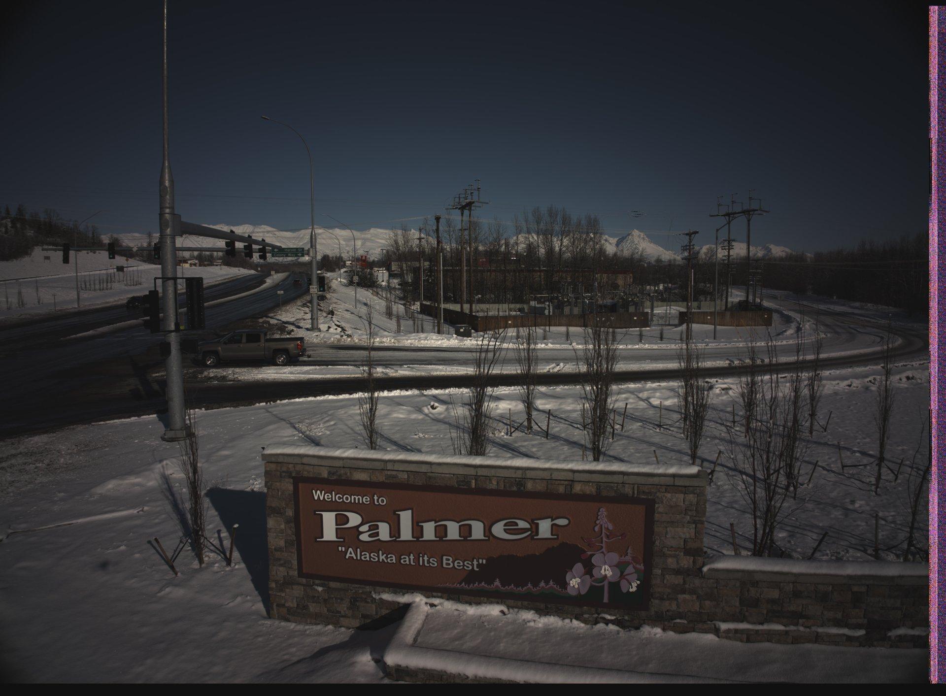 A snowy road intersection in Palmer, Alaska, with a prominent 'Welcome to Palmer' sign in the foreground, snow-covered trees, and distant snow-capped mountains