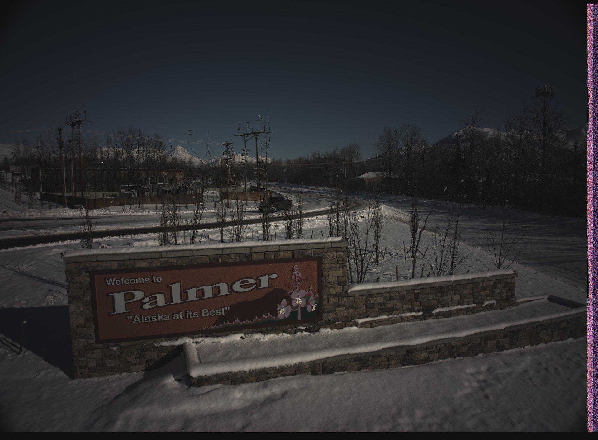 A snowy welcome sign for Palmer, Alaska, set against a backdrop of snow-covered mountains and a winding road with a single truck.