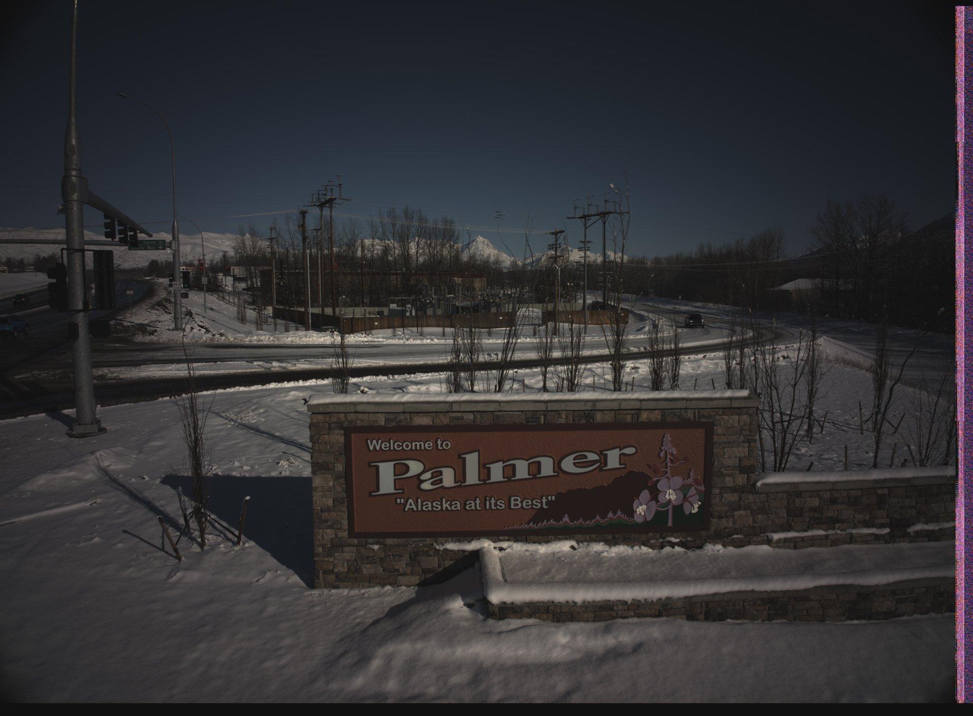 A snowy roadside welcome sign for Palmer, Alaska, with a mountain backdrop and traffic infrastructure under a clear blue sky.