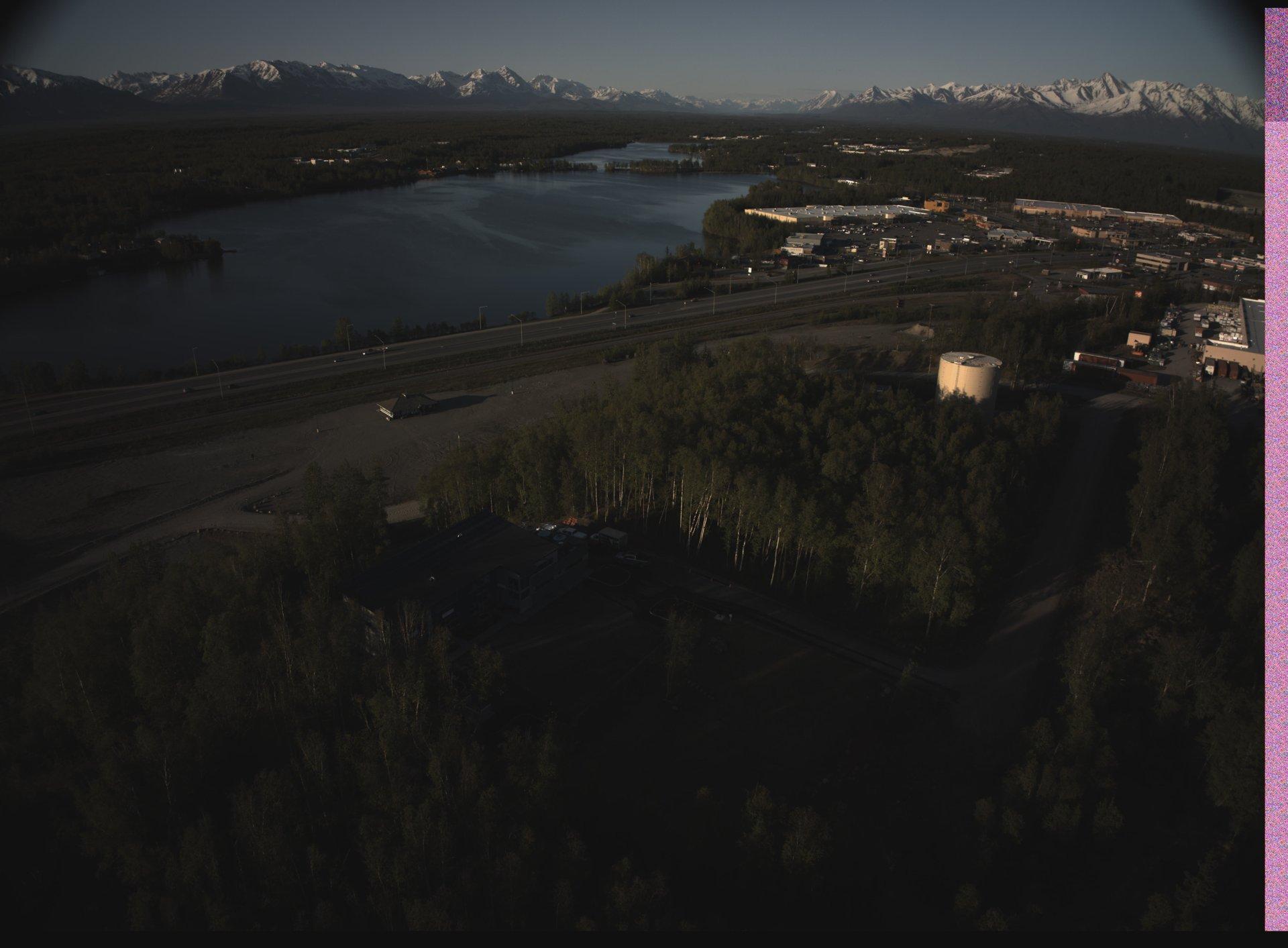 Aerial view of a large lake bordered by forested land and a highway, with snow-capped mountains in the distance and industrial buildings on the far side.